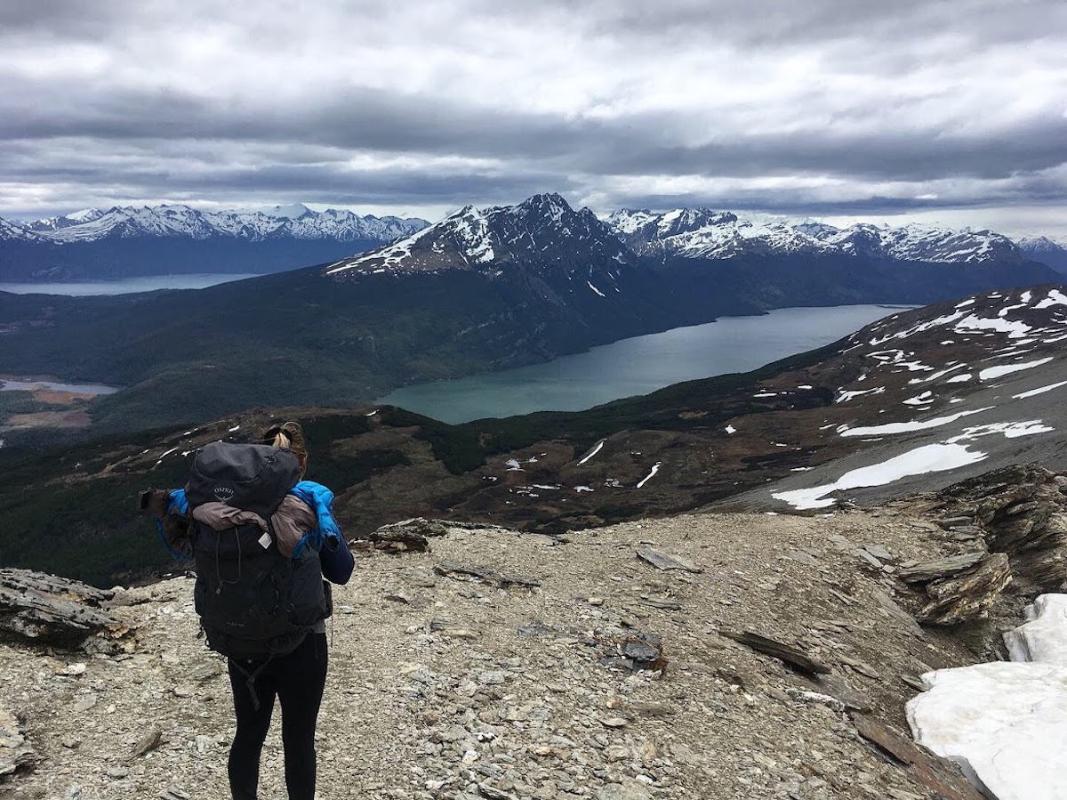 A hiker facing away from the camera with full pack on and overlooking a stunning mountain vistas with light snow dusting the peaks and lakes below. Taken at Cerro Guanaco inTierra del Fuego, Argentina, one of the best day hikes in Patagonia.